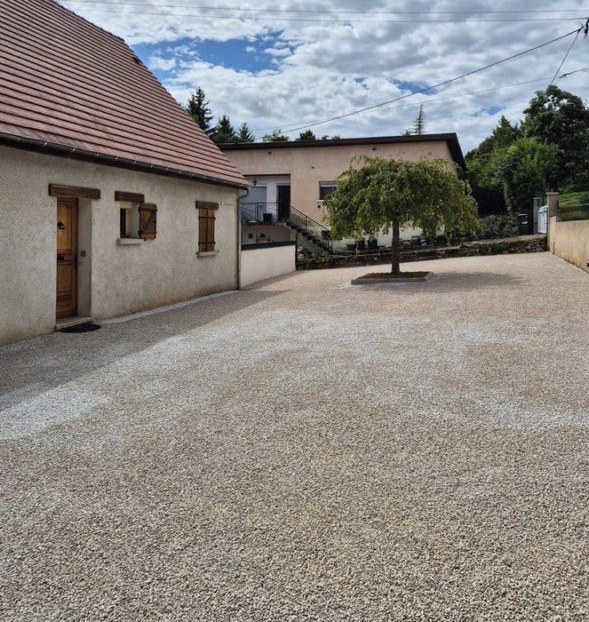 Allée de jardin en gravier avec bordures enherbées, située devant une maison avec volets bois